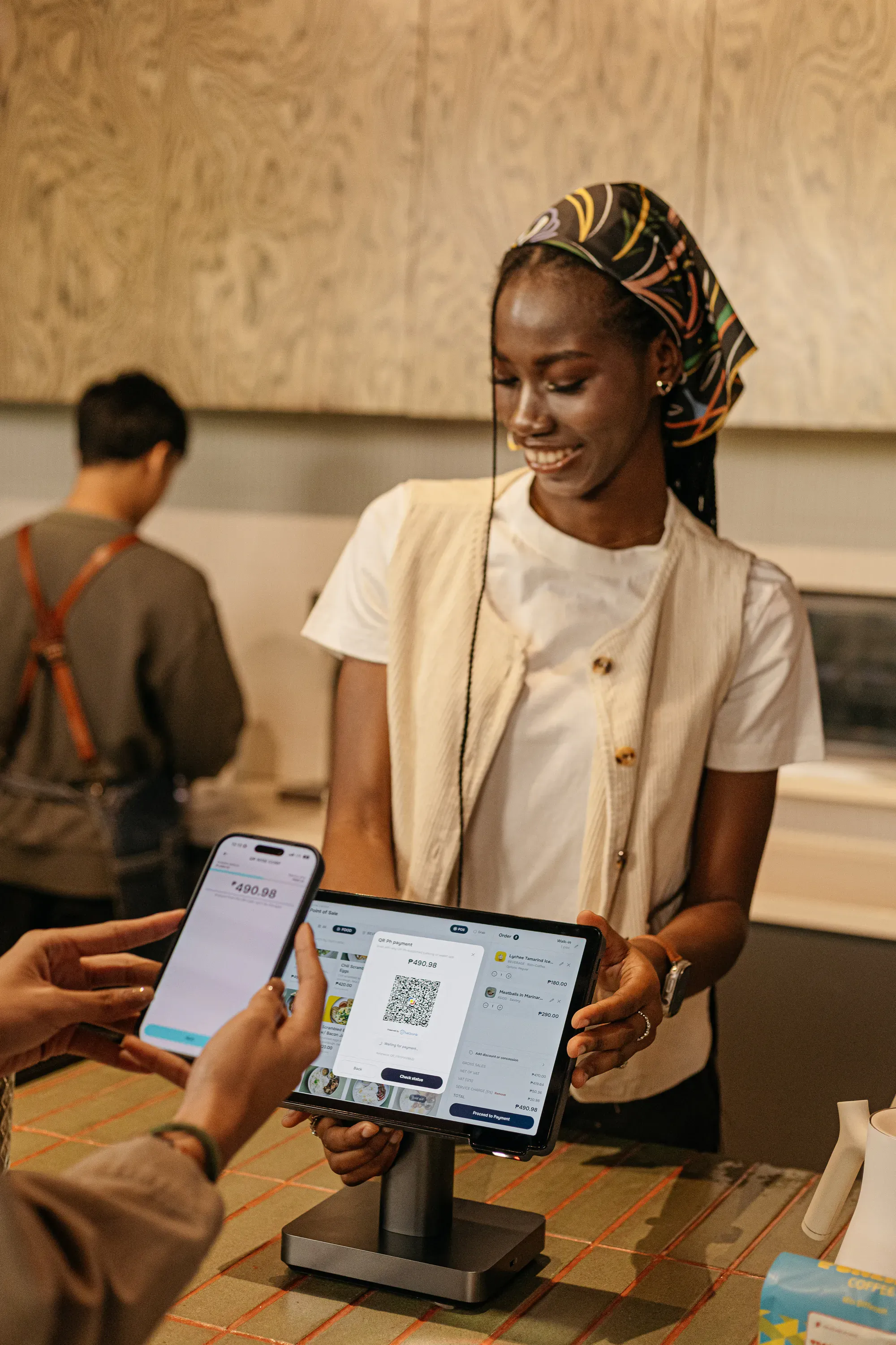 A barista smiling as a customer pays by phone at a counter terminal